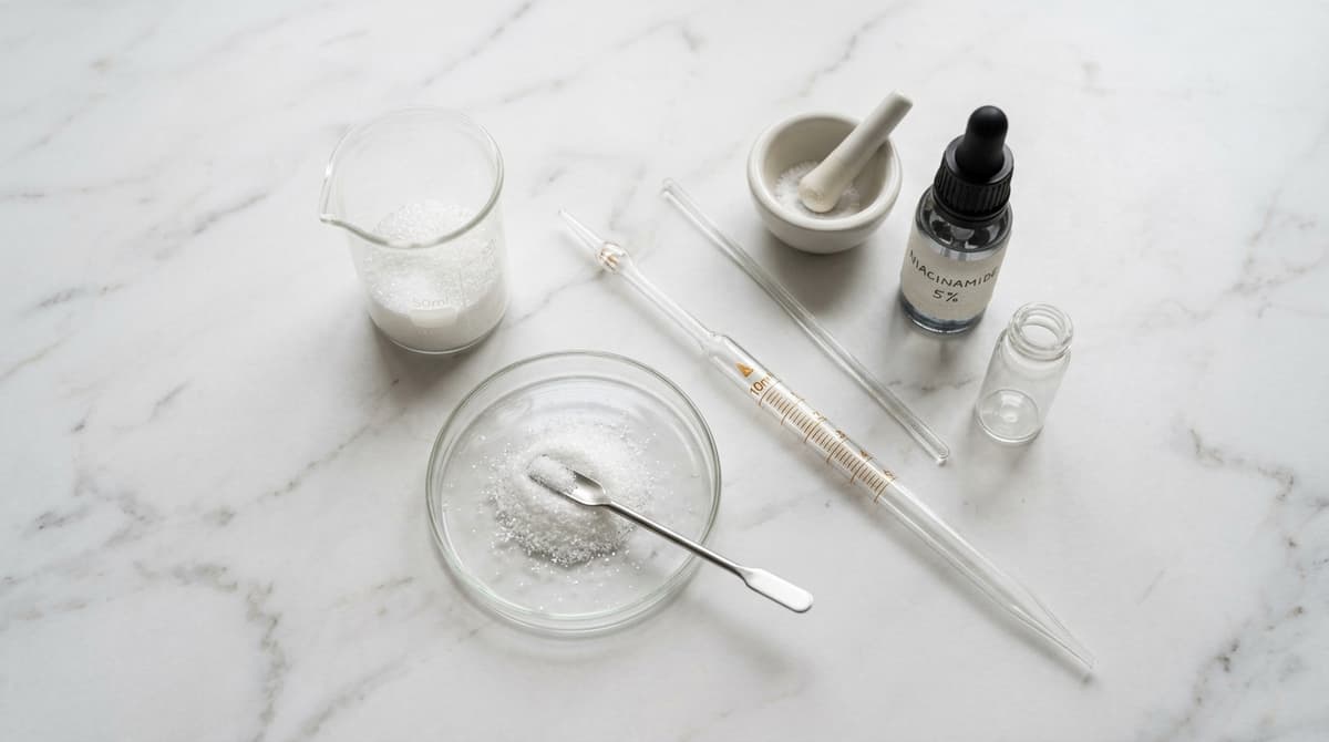 Foto flat lay peralatan gelas laboratorium dan pipet dengan bubuk kristal putih (niacinamide) di atas permukaan marmer, gaya fotografi kecantikan editorial, cahaya alami yang lembut, komposisi minimalis