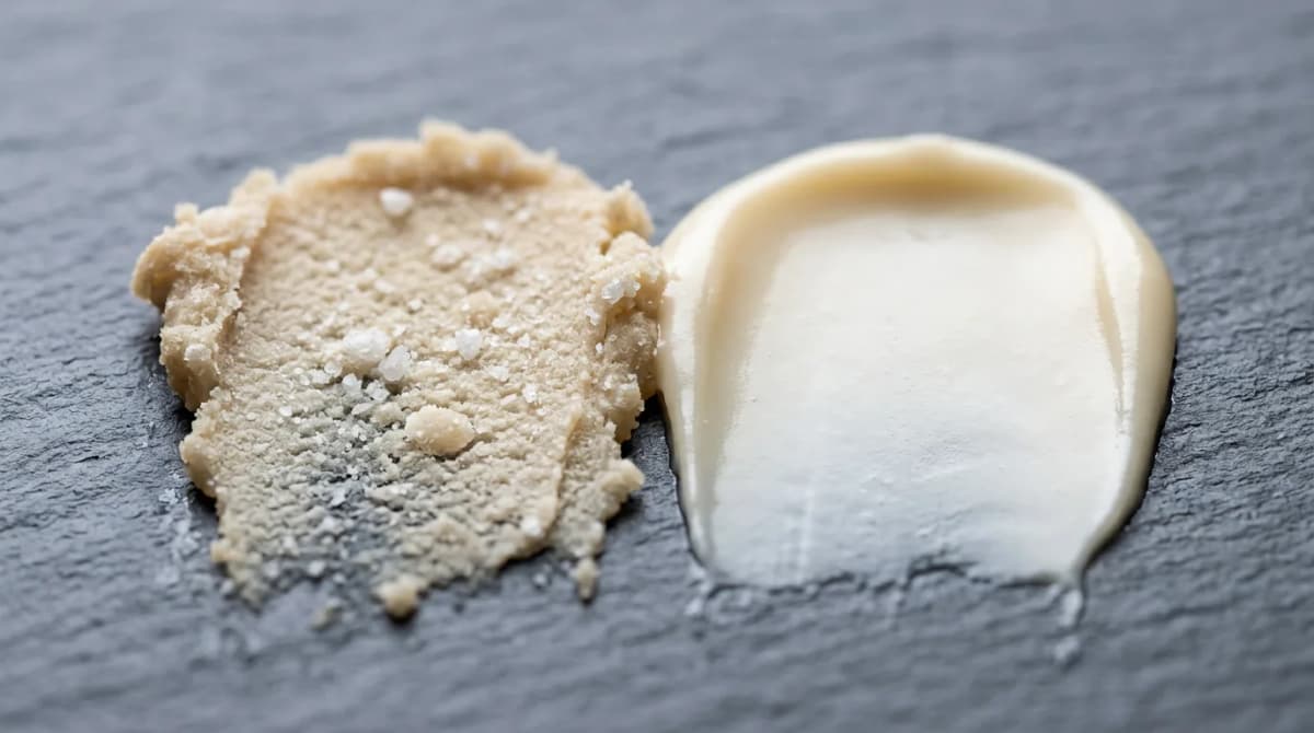 Foto makro dua olesan shea butter pada lempeng batu abu-abu: olesan di sebelah kiri berpasir dengan kristal keras yang terlihat, sedangkan olesan di sebelah kanan sangat halus dan berkilau